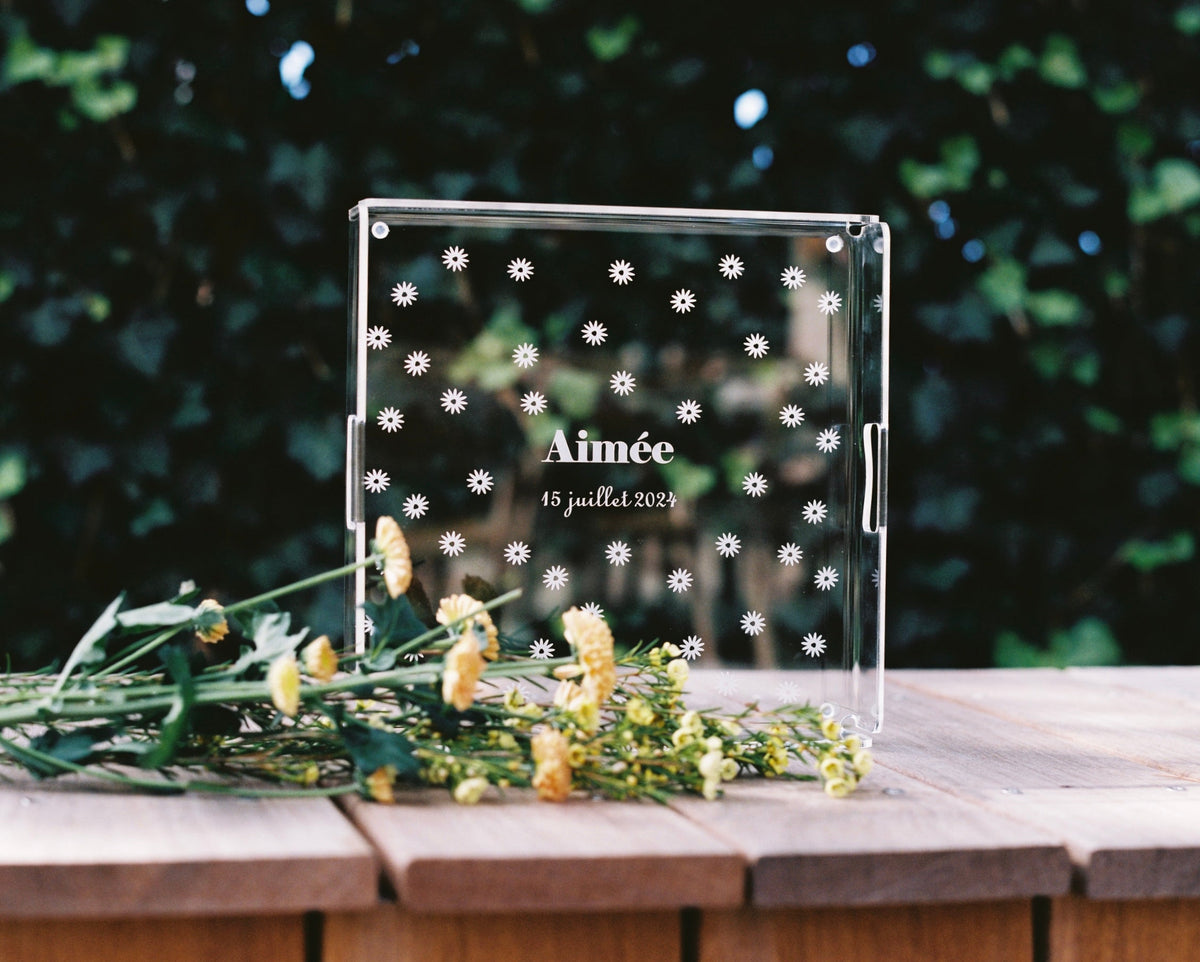 Plateau carré gravé « Aimée, 15 juillet 2024 » semé de marguerites, sur une table de jardin en bois