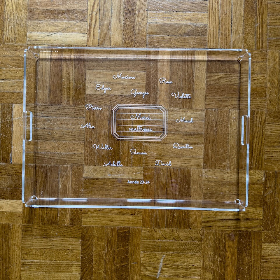 Plateau rectangulaire gravé « Merci maîtresse » entouré des prénoms de la classe, sur un parquet en point de Hongrie