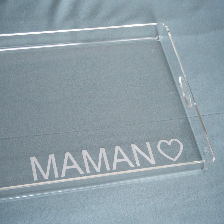 Gros plan sur l'angle d'un plateau en plexi gravé « MAMAN ♡ » en capitales, sur un tissu bleu-gris