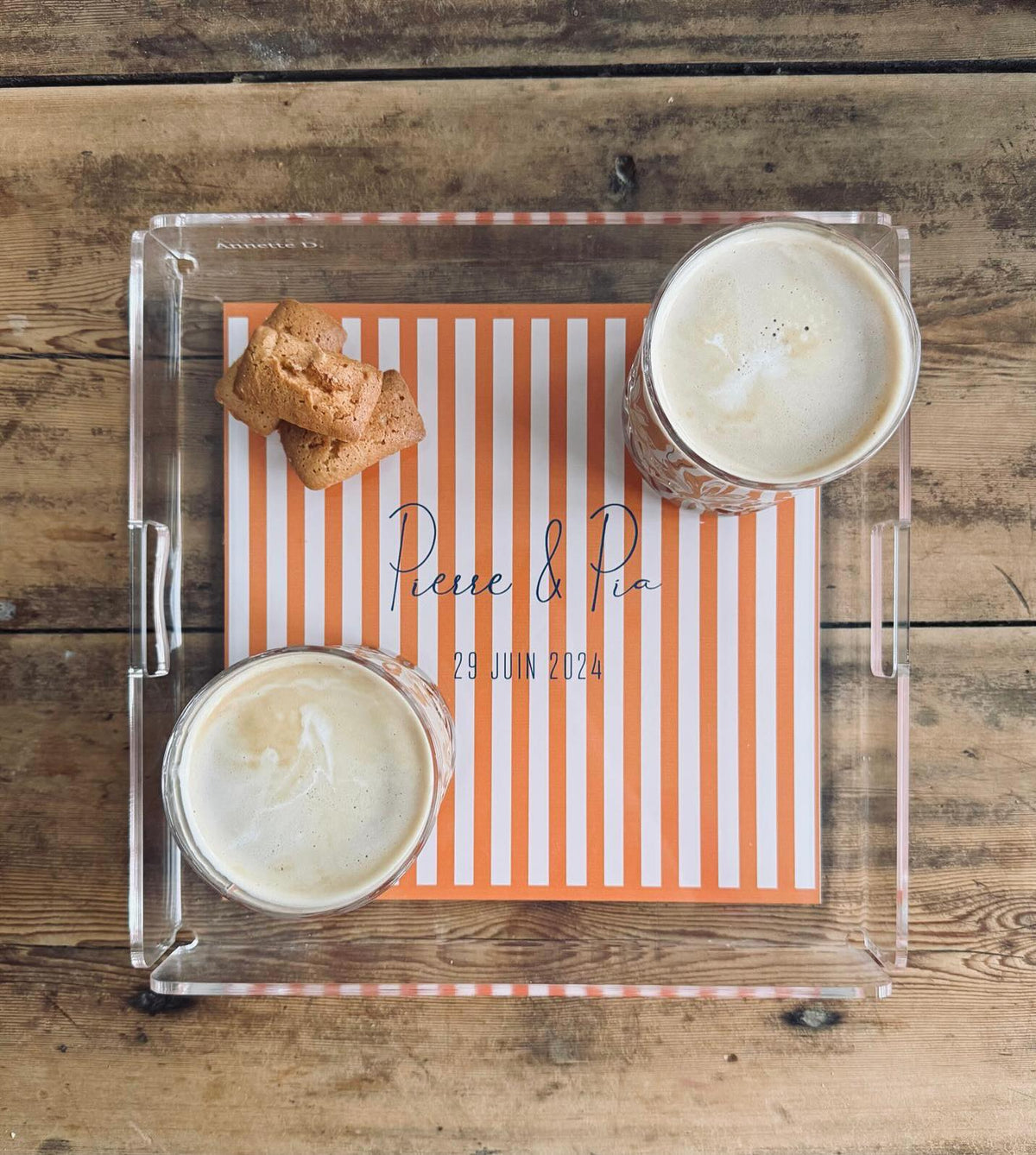 Plateau en plexi à rayures orange marqué « Pierre & Pia, 29 juin 2024 », deux cafés et des biscuits posés dessus, sur une table en bois
