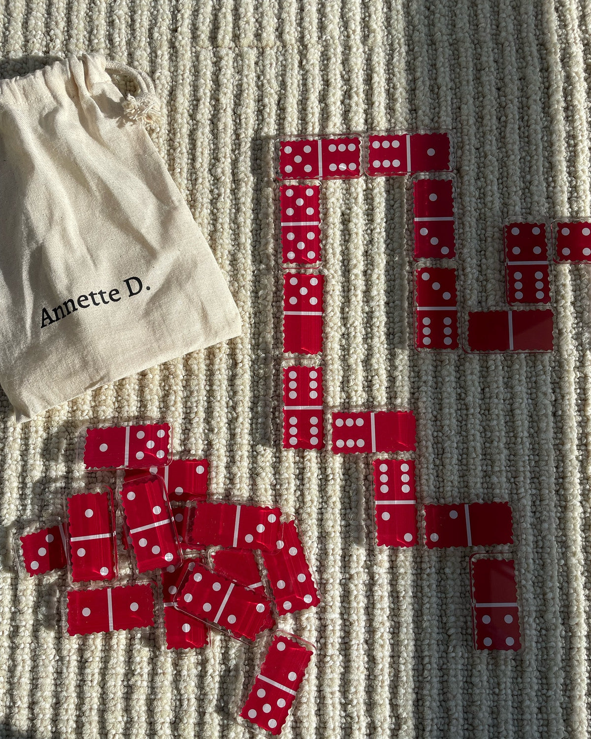 Dominos rouges en plexi étalés sur un tapis, avec leur pochette en tissu Annette D.