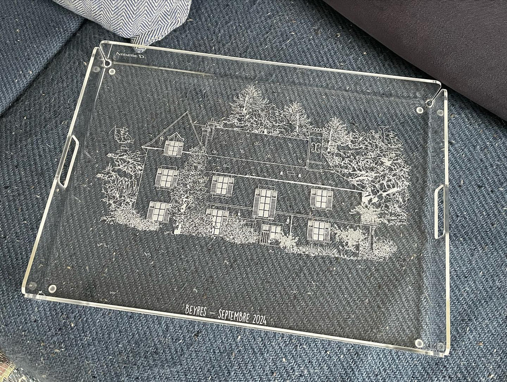 Plateau gravé d’une maison entourée d’arbres, posé sur un tissu gris