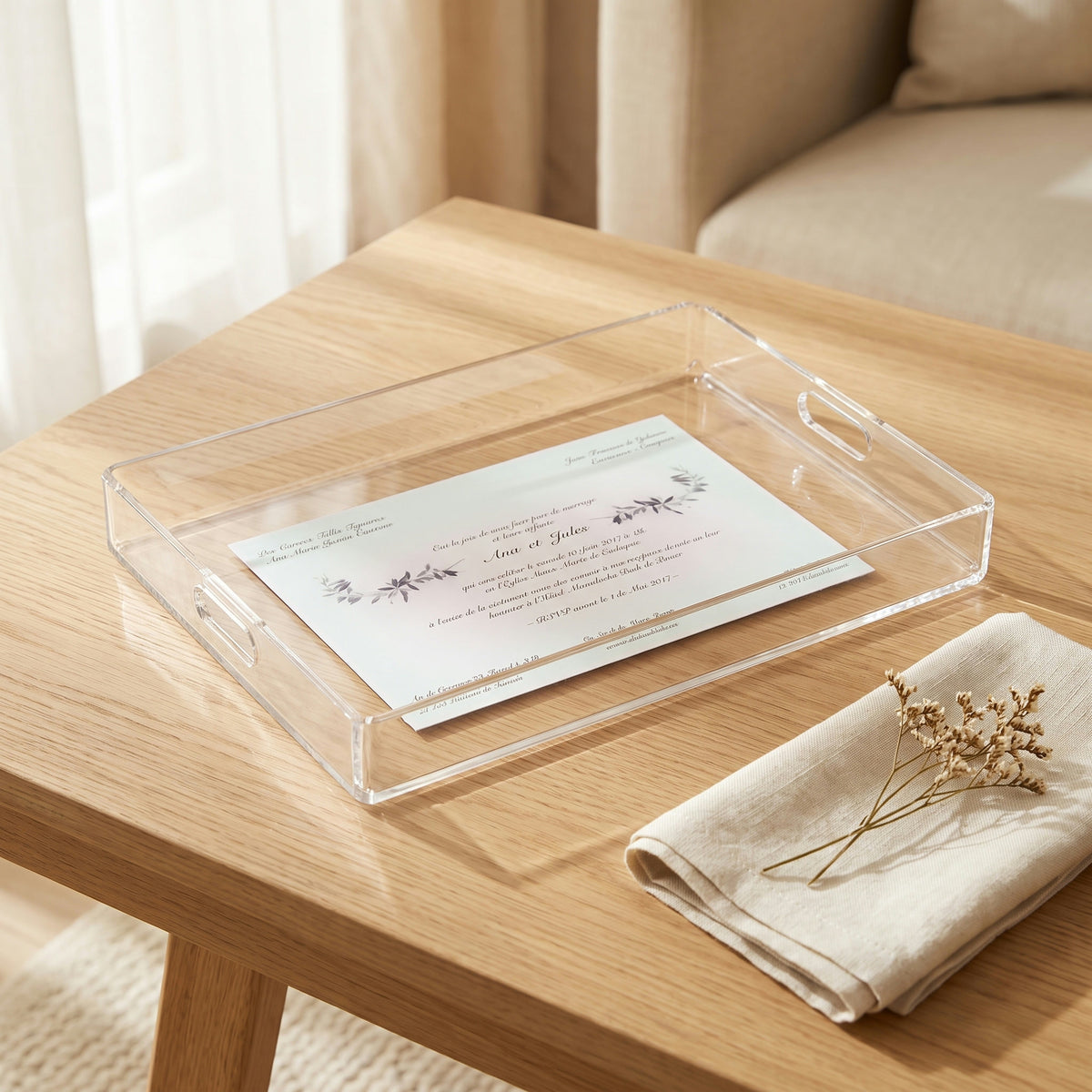 Plateau en plexi avec un faire-part de mariage imprimé, posé sur une table en chêne clair près d'une fenêtre, à côté d'une serviette en lin et d'un brin de fleurs séchées
