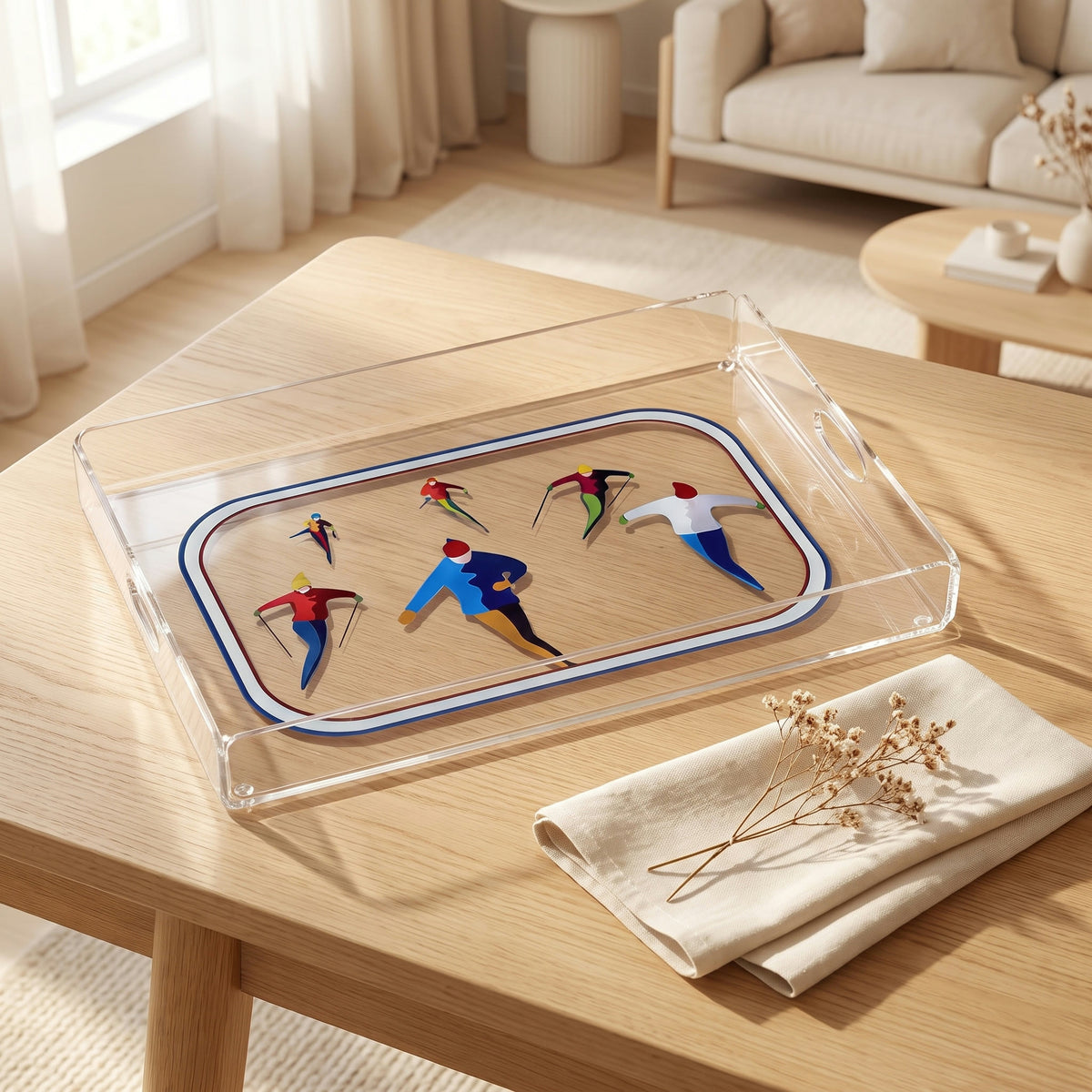 Plateau en plexi transparent imprimé de six skieurs bleu, blanc et rouge, posé sur une table en chêne clair avec une serviette en lin et des fleurs séchées