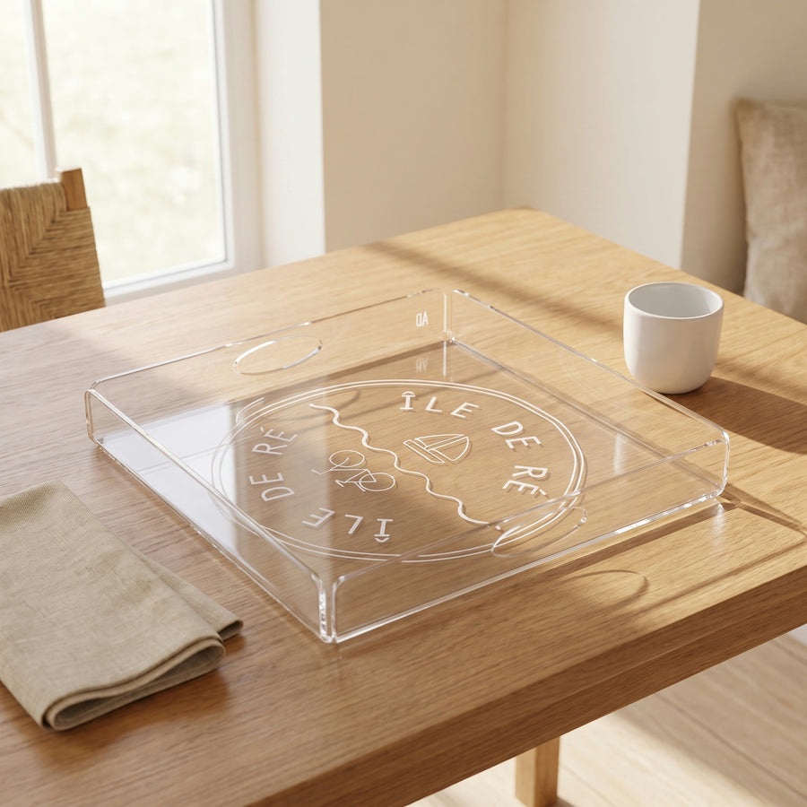 Plateau carré en plexi gravé « Île de Ré », avec un vélo et un voilier, posé sur une table en chêne à côté d'une tasse blanche et d'une serviette en lin