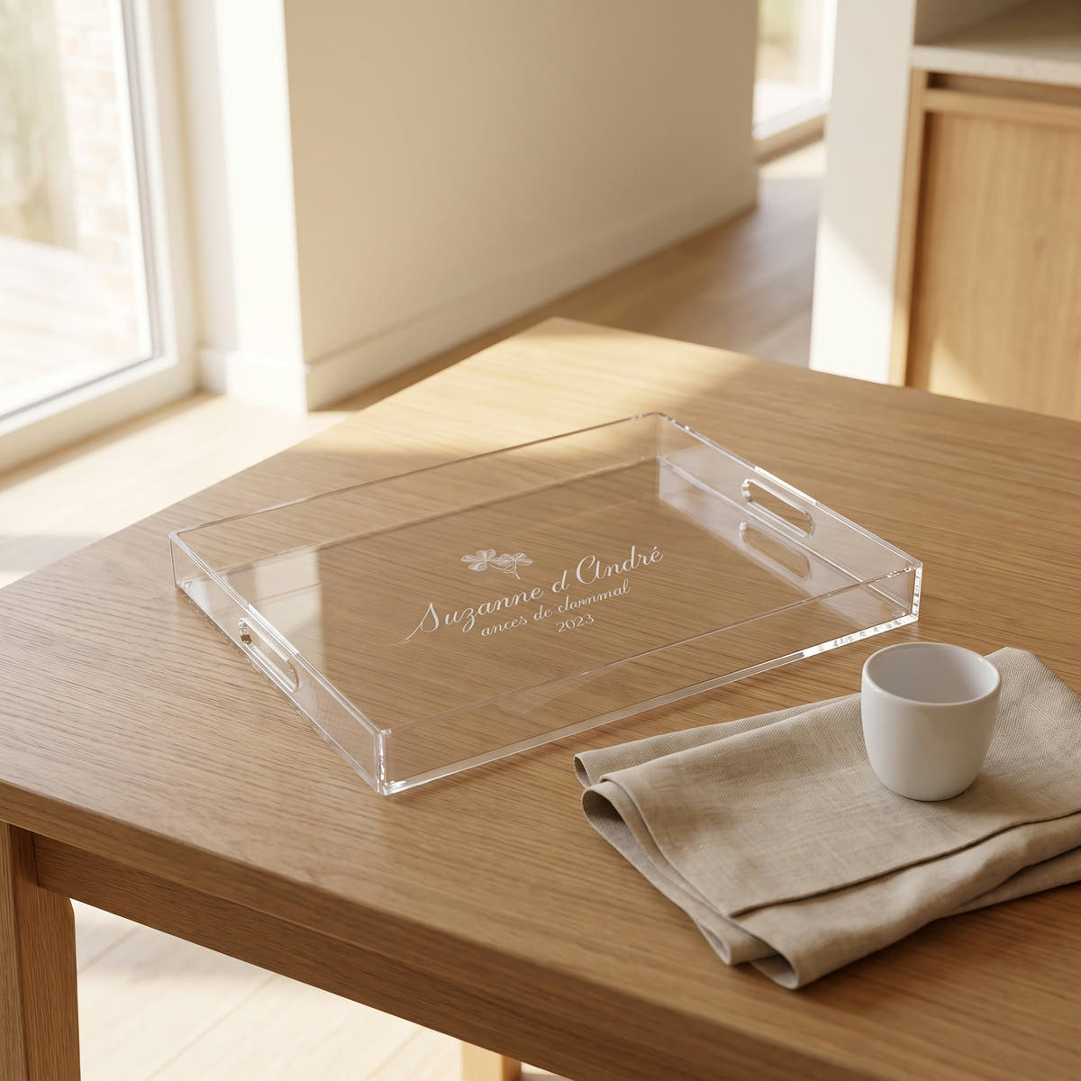 Plateau en plexi gravé des prénoms des mariés, posé sur une table en chêne près d'une fenêtre, avec une tasse blanche et une serviette en lin