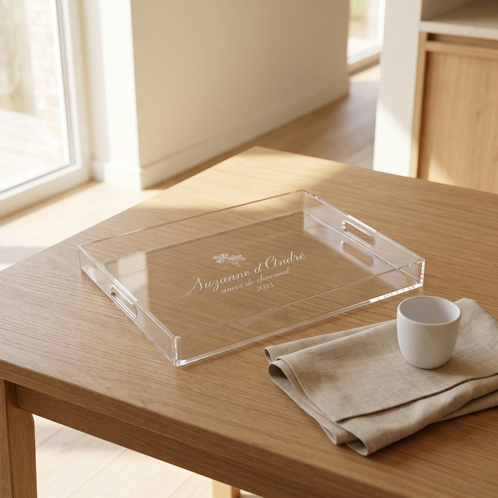 Plateau en plexi gravé des prénoms des mariés, posé sur une table en chêne près d'une fenêtre, avec une tasse blanche et une serviette en lin