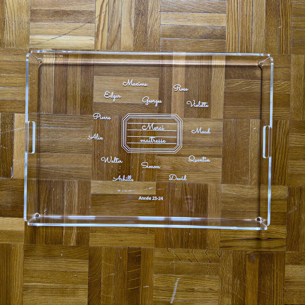 Plateau rectangulaire gravé « Merci maîtresse » entouré des prénoms de la classe, sur un parquet en point de Hongrie
