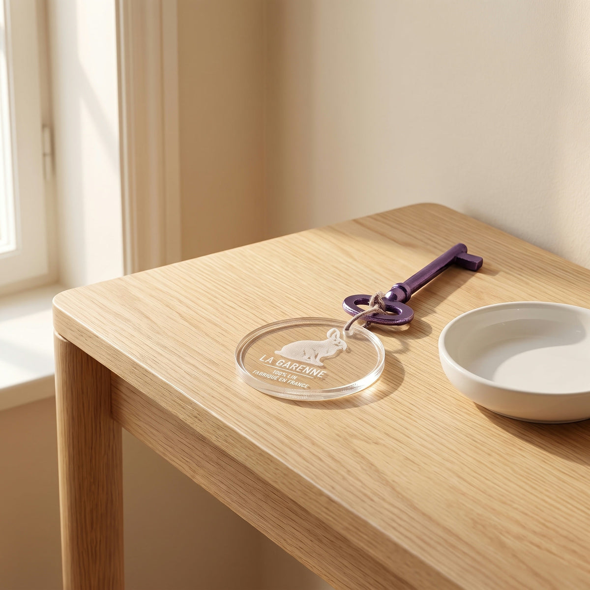 Porte-clés rond en plexi gravé d'un lapin et de l'inscription « La Garenne », attaché à une ancienne clé violette, sur une console en chêne près d'une coupelle blanche