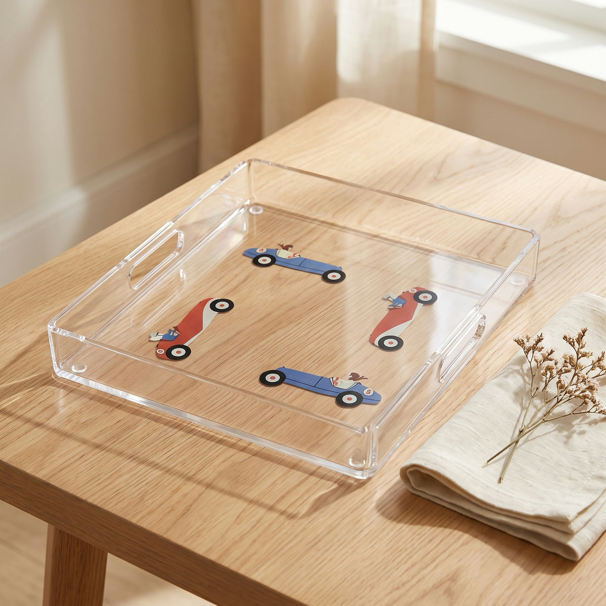 Plateau en plexi imprimé de quatre voitures de course anciennes bleues et rouges, posé sur une table en chêne clair avec une serviette en lin et des fleurs séchées